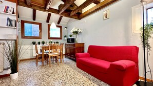 Un salon chaleureux avec un canapé rouge, une table à manger en bois et des chaises. On y trouve une bibliothèque, une plante en pot et une fenêtre donnant sur un beau paysage.