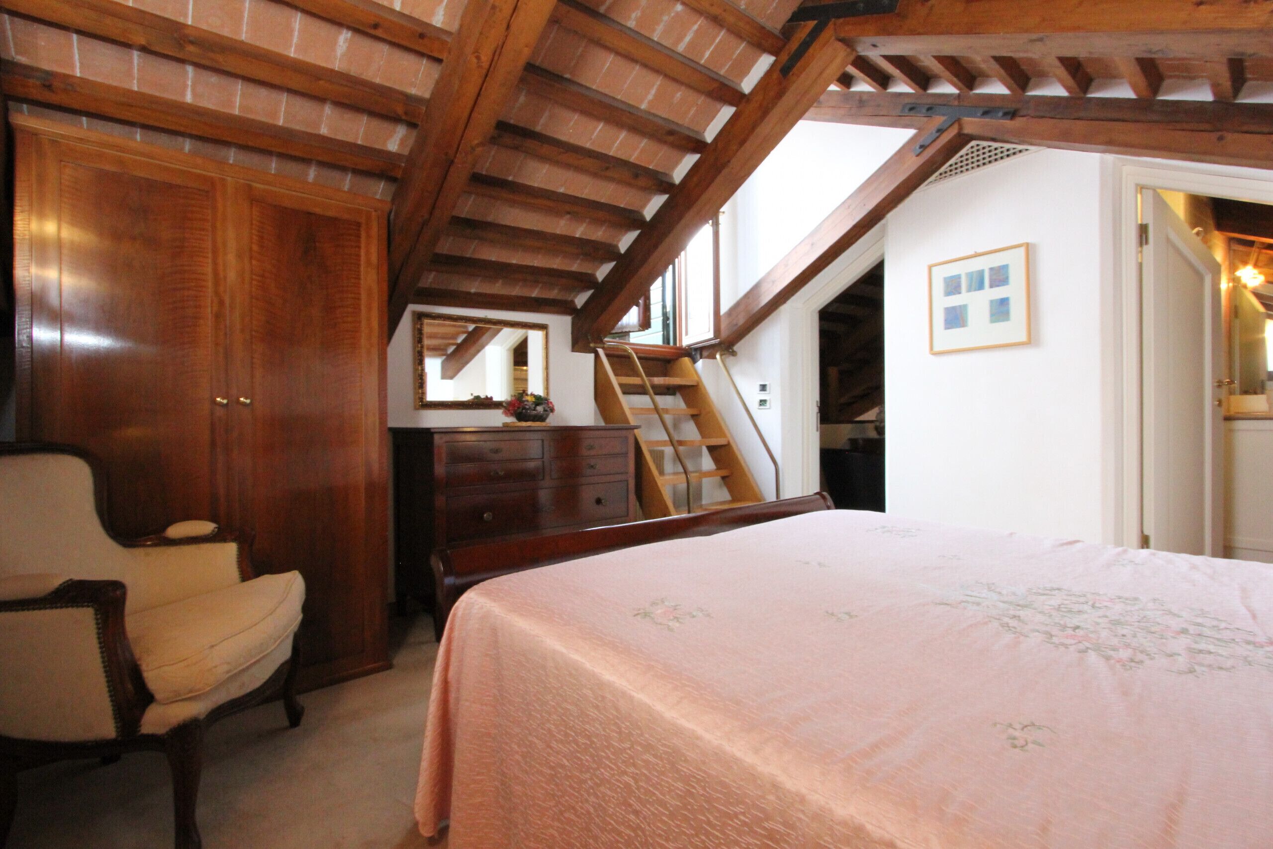 A bedroom with a wooden ceiling, a bed with a floral bedspread, a chair, a dresser, and a mirror on the wall.
