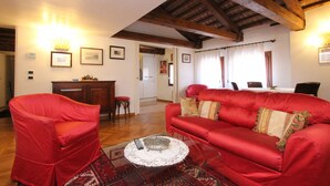 Un salon avec un canapé rouge, un fauteuil rouge, une table basse ornée d’un chemin de table en dentelle et des poutres en bois au plafond.