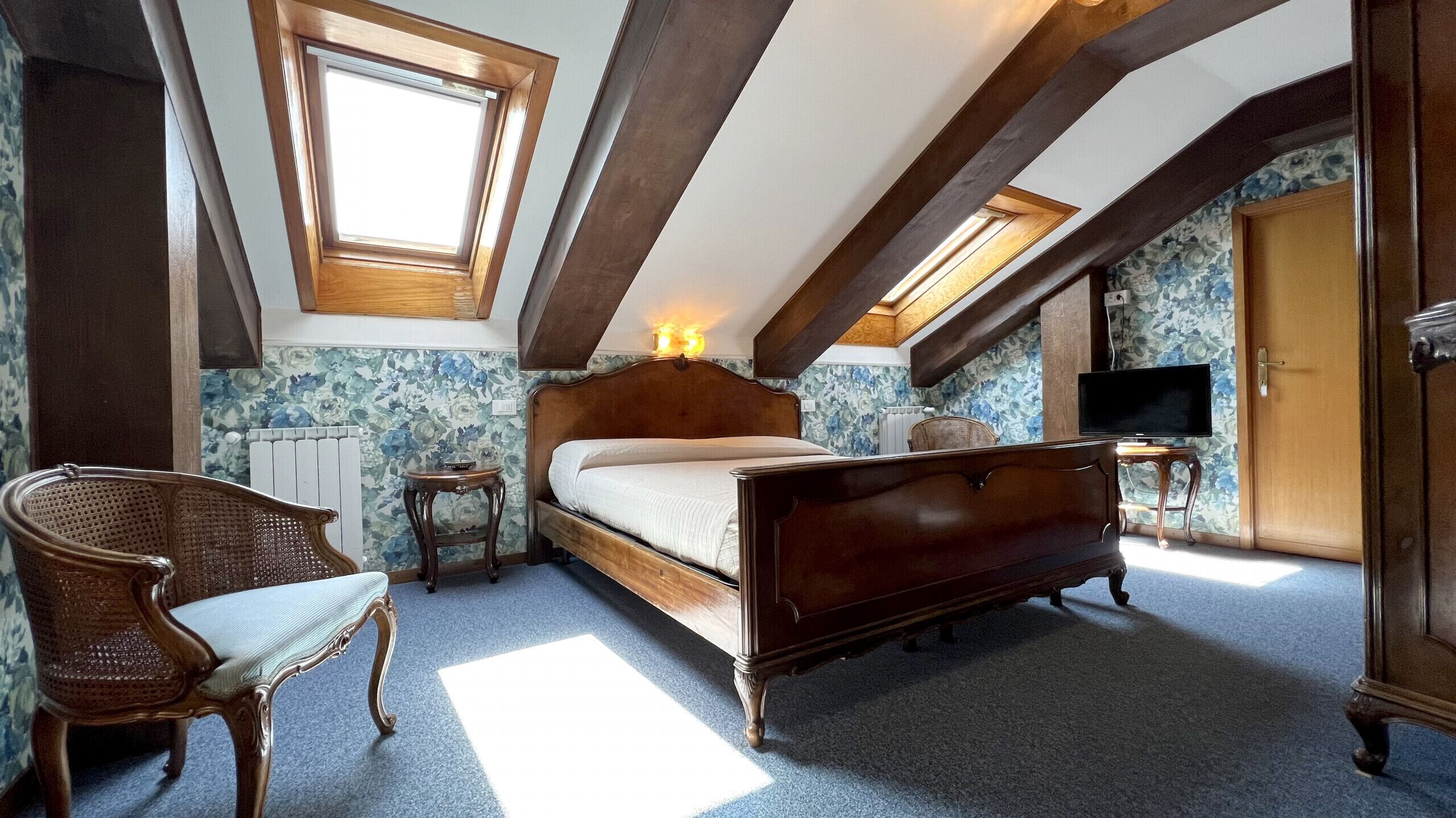 A bedroom with a wooden bed, a chair, a small table, a TV, and a window.