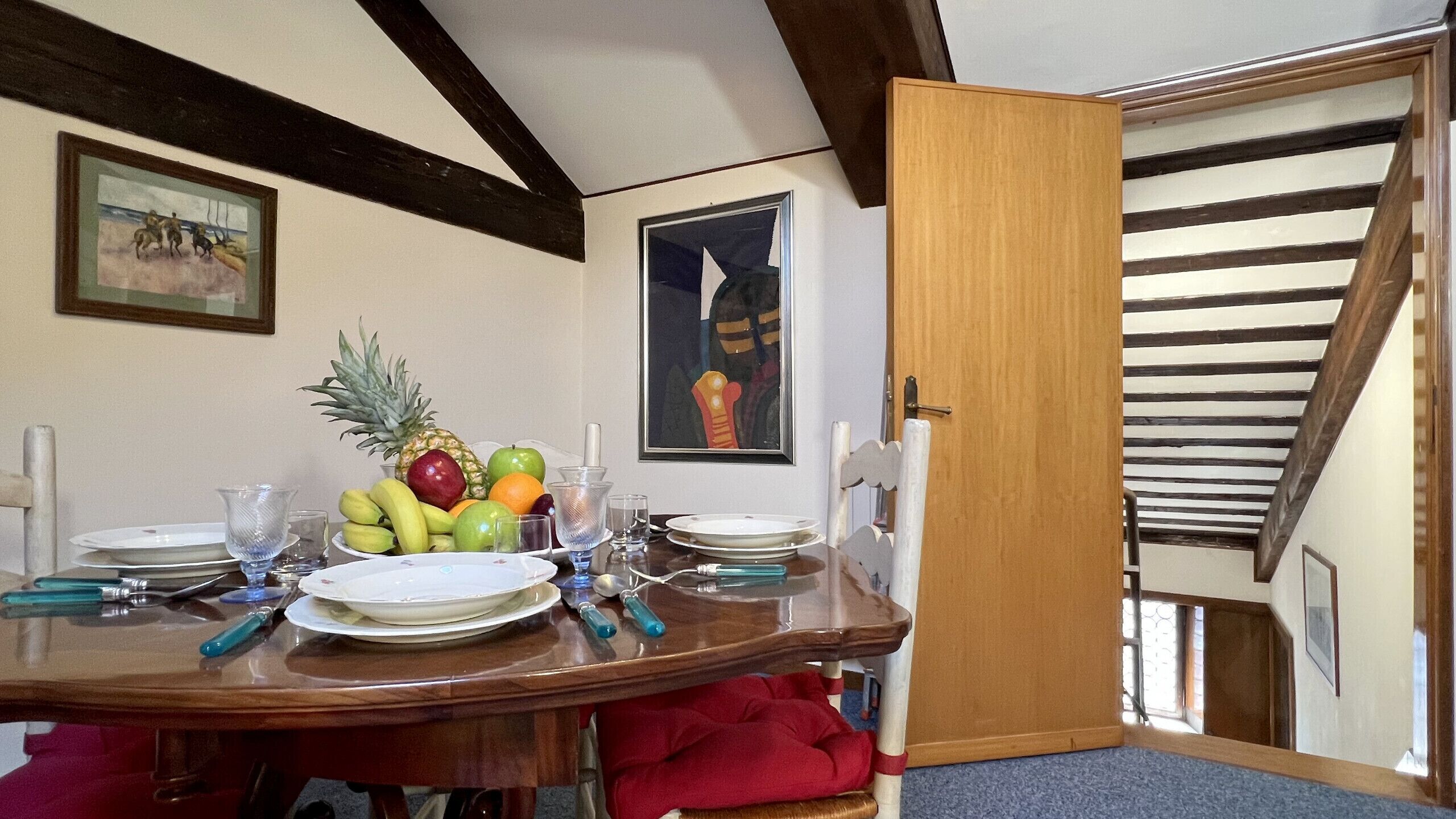 A dining area with a wooden table set for four, fruit on the table, and a painting on the wall.