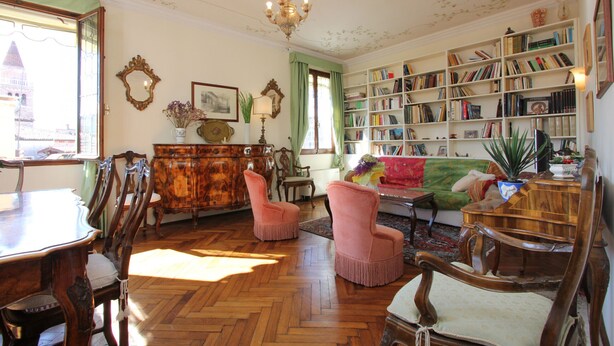 Um quarto com piso de madeira em padrão herringbone, uma grande estante de livros, uma mesa de jantar com cadeiras e um sofá.