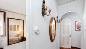 A hallway with a bed, a wall-mounted thermostat, a mirror, a candelabra, and a door with a white frame.