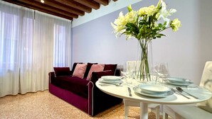 A dining area with a round table set for four, a dark red sofa, and a vase of white flowers. - San Marco Canal View (200m from St. Mark´s Square) (Venecia)