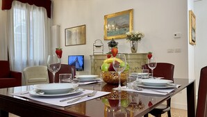 A dining room with a table set for four, including plates, glasses, and cutlery. A fruit bowl with bananas, apples, and grapes is on the table. There are two chairs, a sofa, and a television in the background. - Manin Terrace (200m from St Mark´s Square) (Venecia)