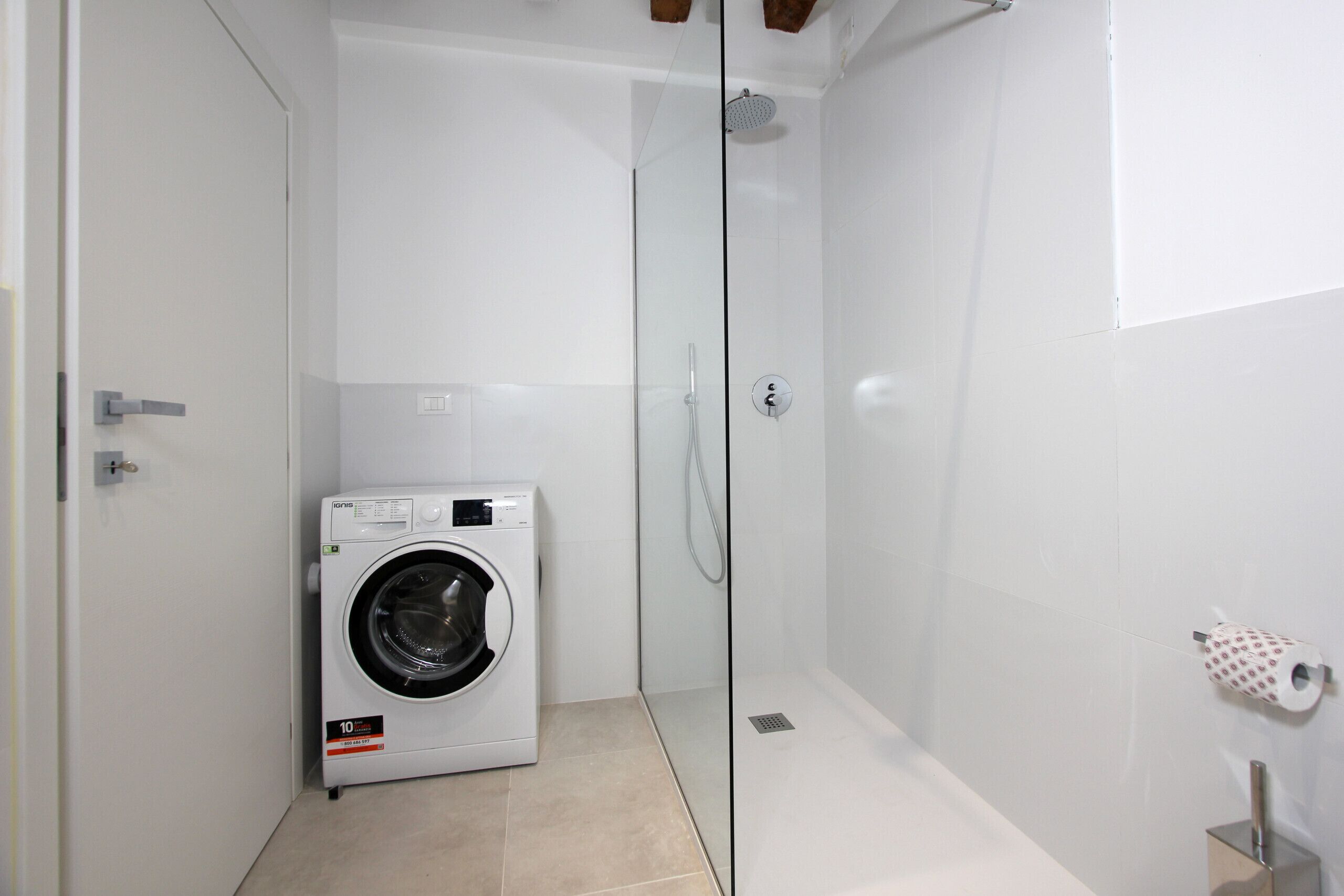 Une petite buanderie équipée d’un lave-linge à chargement frontal, d’une douche à parois de verre et d’un porte-rouleau de papier toilette fixé au mur.