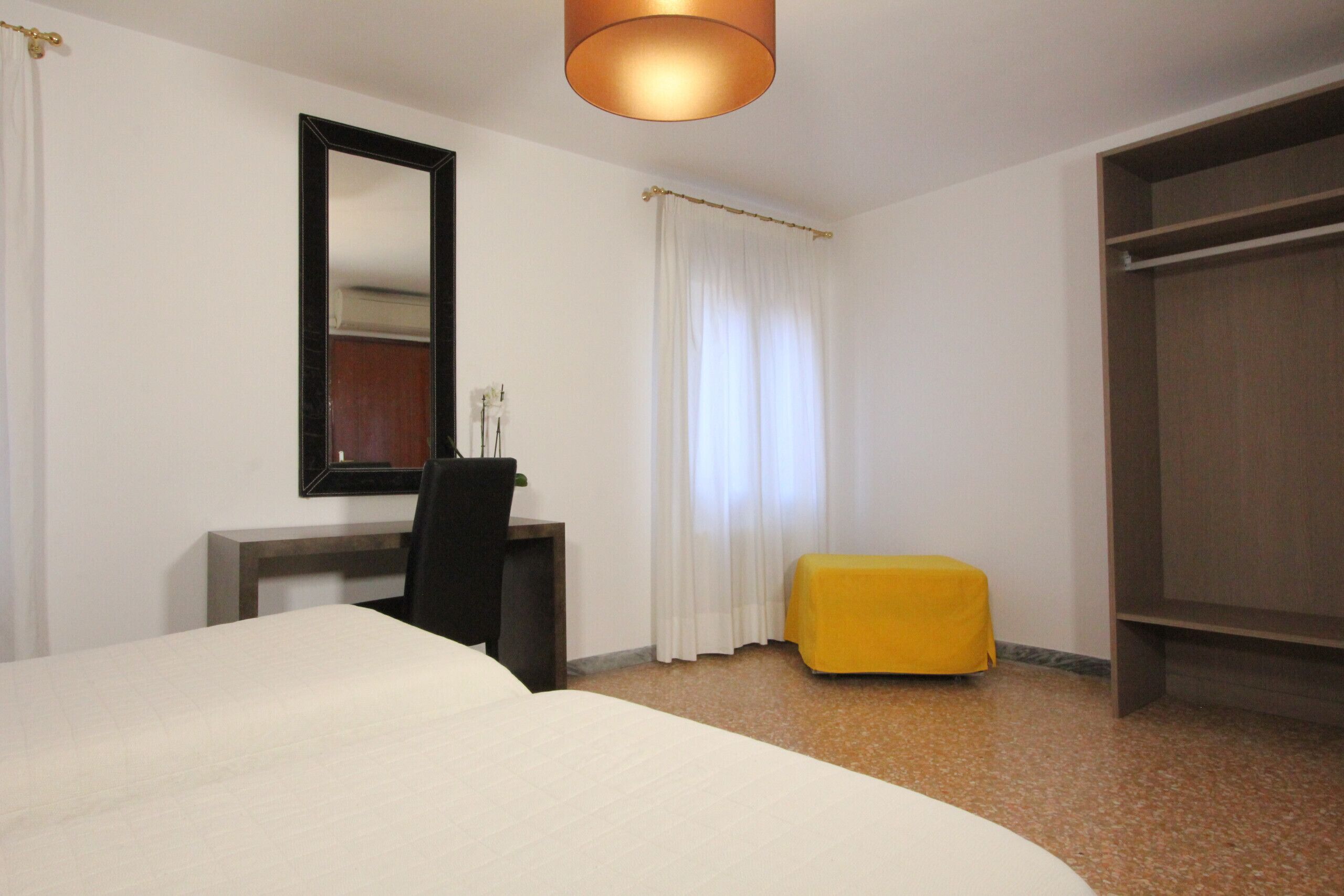 A hotel room with a bed, a desk, a chair, a small yellow ottoman, a wardrobe, and a mirror on the wall.