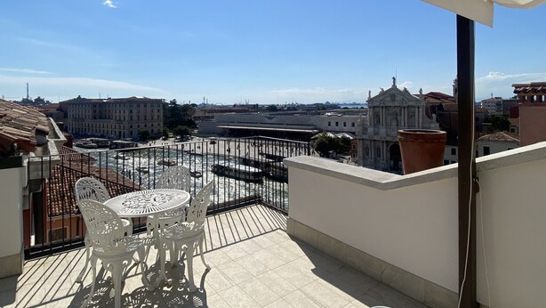 En balkon med et bord og stole, med udsigt over en by med historiske bygninger og en havn.