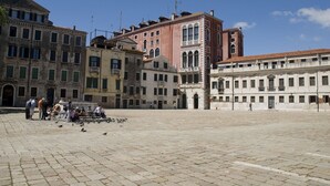 Una plaza histórica con edificios de arquitectura clásica, un espacio abierto en el centro y palomas en el suelo.