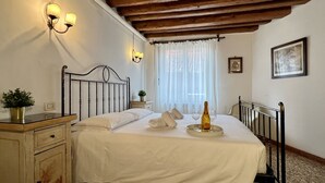Ein Schlafzimmer mit Bett, Nachttischlampen, einem Nachttisch, einem Blumentopf, einem Fenster mit Vorhängen und einem gerahmten Bild an der Wand.