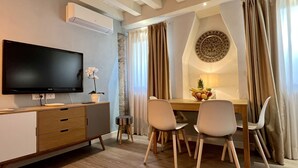 A hotel room with a flat-screen TV, a wooden desk with drawers, a small potted orchid, a dining table with two chairs, a fruit bowl, and a decorative wall piece. - Acquamarina Terrace (Historical Center) (Venecia)