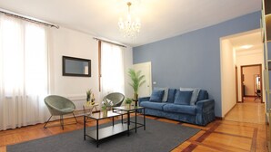 A living room with a blue sofa, two green chairs, a glass coffee table, a chandelier, and a wooden floor.