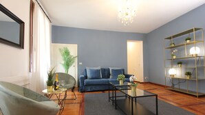 A living room with a blue sofa, a glass coffee table, a green armchair, and a gold shelf with plants.
