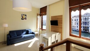A modern living room with a sofa, dining table, and chairs, a TV mounted on the wall, and a large window with a view of buildings and water. - Apartment Grand Canal View (with lift) (Venecia)