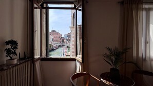 A room with a window overlooking a canal, a chair, a table, and a potted plant. - Apt. Fosca Canal View (Venecia)