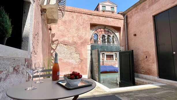 A table with a bottle and a bowl of strawberries, a wine glass, and a view of a courtyard with a pool and a building in the background.