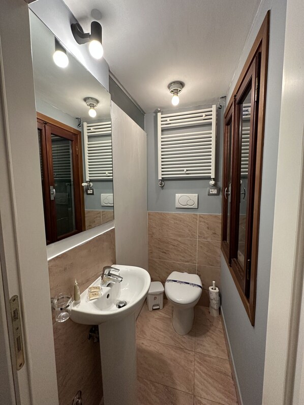 A bathroom with a sink, toilet, and mirror. There are ceiling lights, a window with blinds, and a radiator. - Residence Flowery Terrace (12 pax) (Venecia)