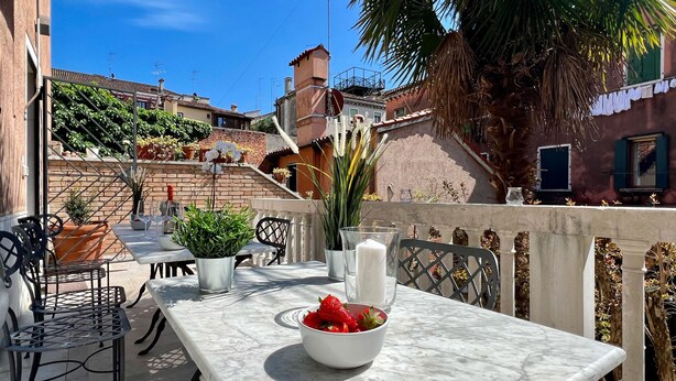 An outdoor terrace with a table set for dining, surrounded by chairs, a bowl of strawberries, and potted plants.