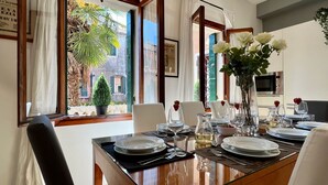 A dining area with a table set for two, a potted plant, and a view of the street outside.