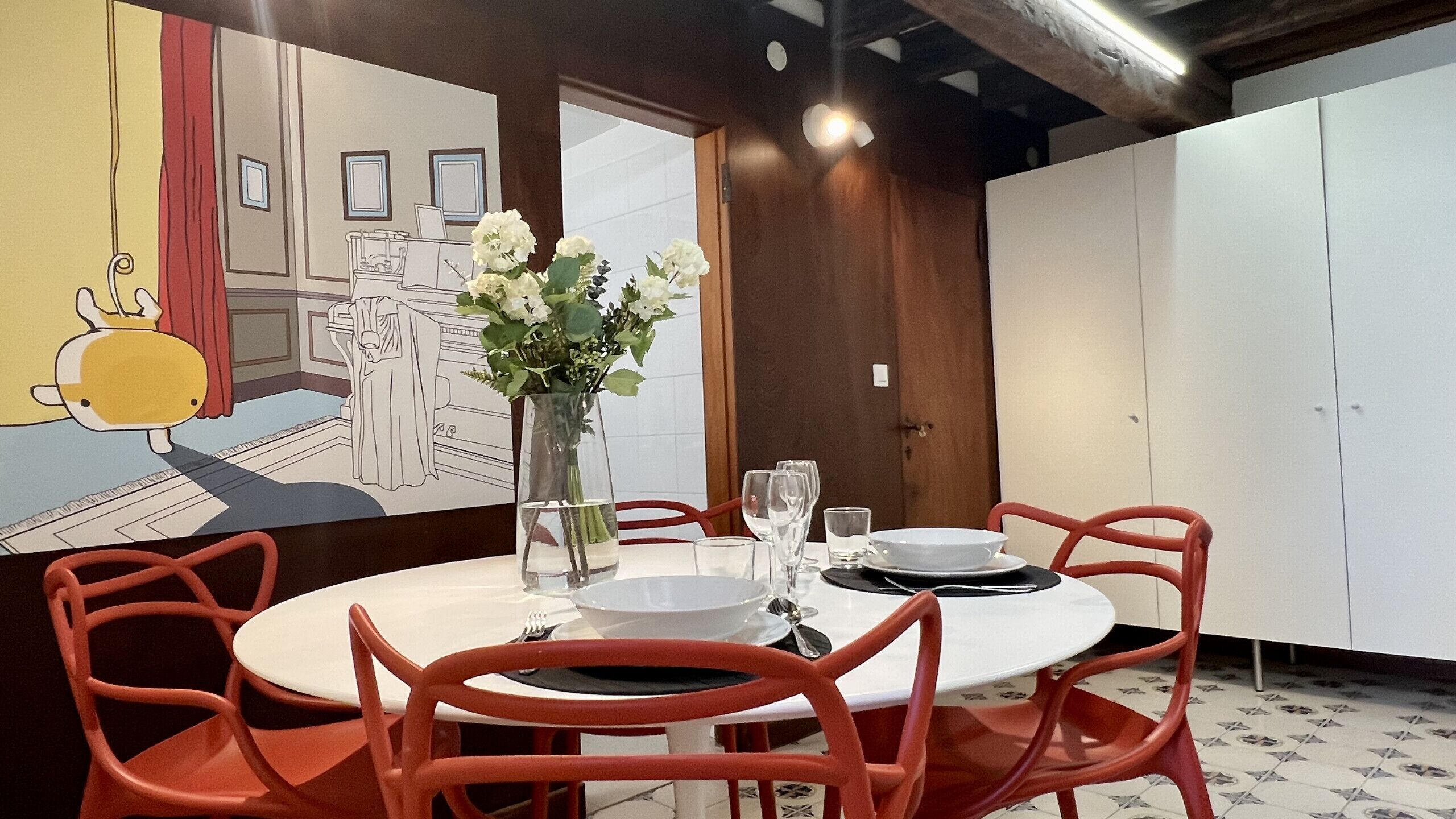 A dining area with a round table set for four, surrounded by red chairs. A vase with white flowers is on the table.