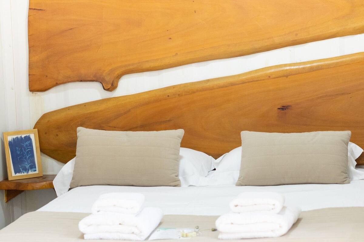 A hotel room with a wooden headboard, two beige pillows, a neatly made bed with white linens, and a framed picture on the nightstand.