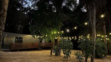 Un espacio exterior rústico con un banco de madera y luces colgantes, rodeado de árboles y un edificio con amplios ventanales.
