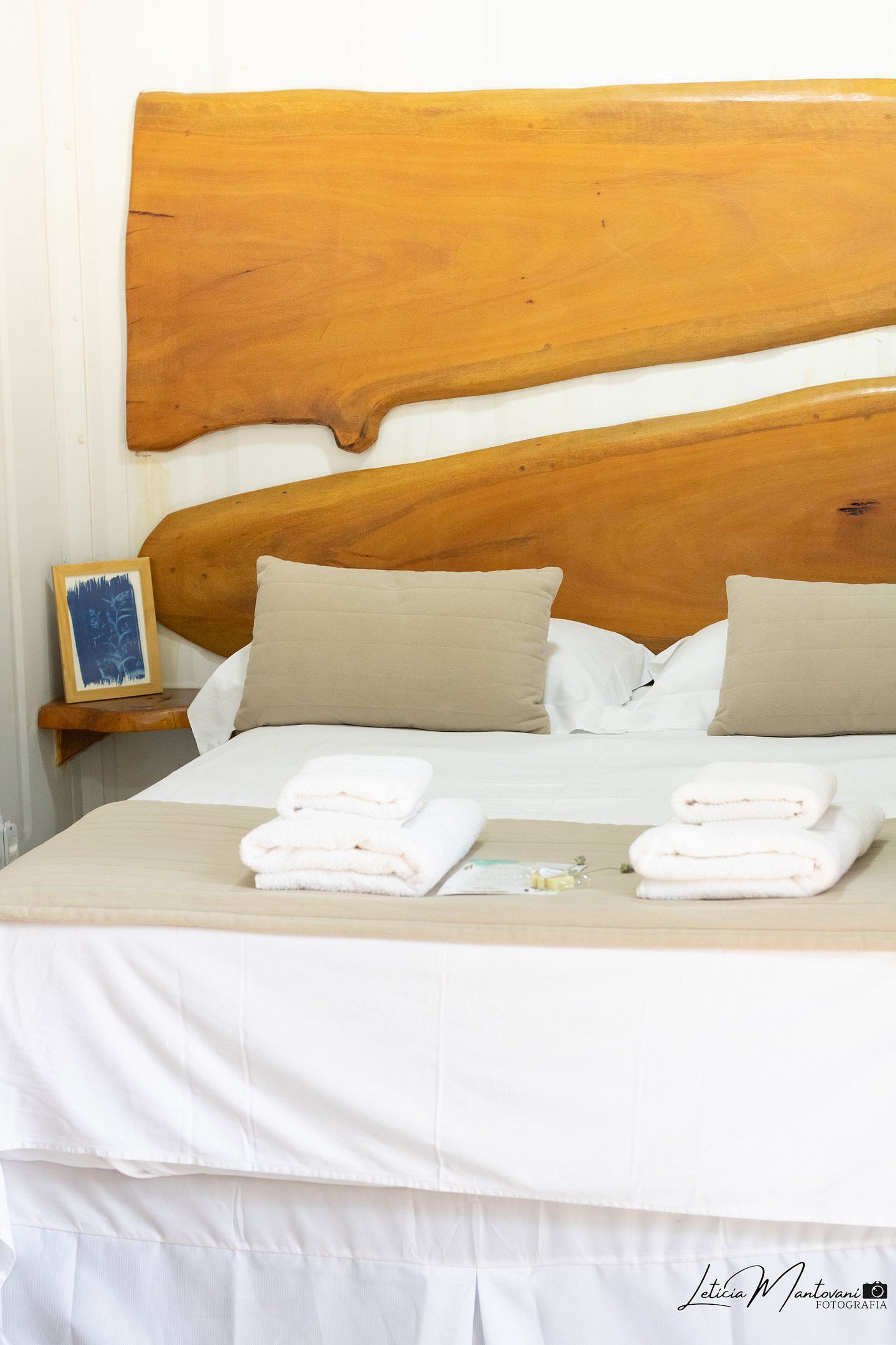 Una cama bien tendida con sábanas blancas y dos almohadas color beige, un cabecero de madera y una mesita de noche con un cuadro enmarcado.