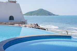 A swimming pool with clear blue water, a white building with a window, and a view of the sea and a green hill in the background.