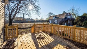 Een houten terras met een balustrade, een boom en een huis op de achtergrond.