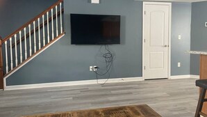 Un salon avec un mur gris, un téléviseur fixé au mur, une porte blanche et un escalier.