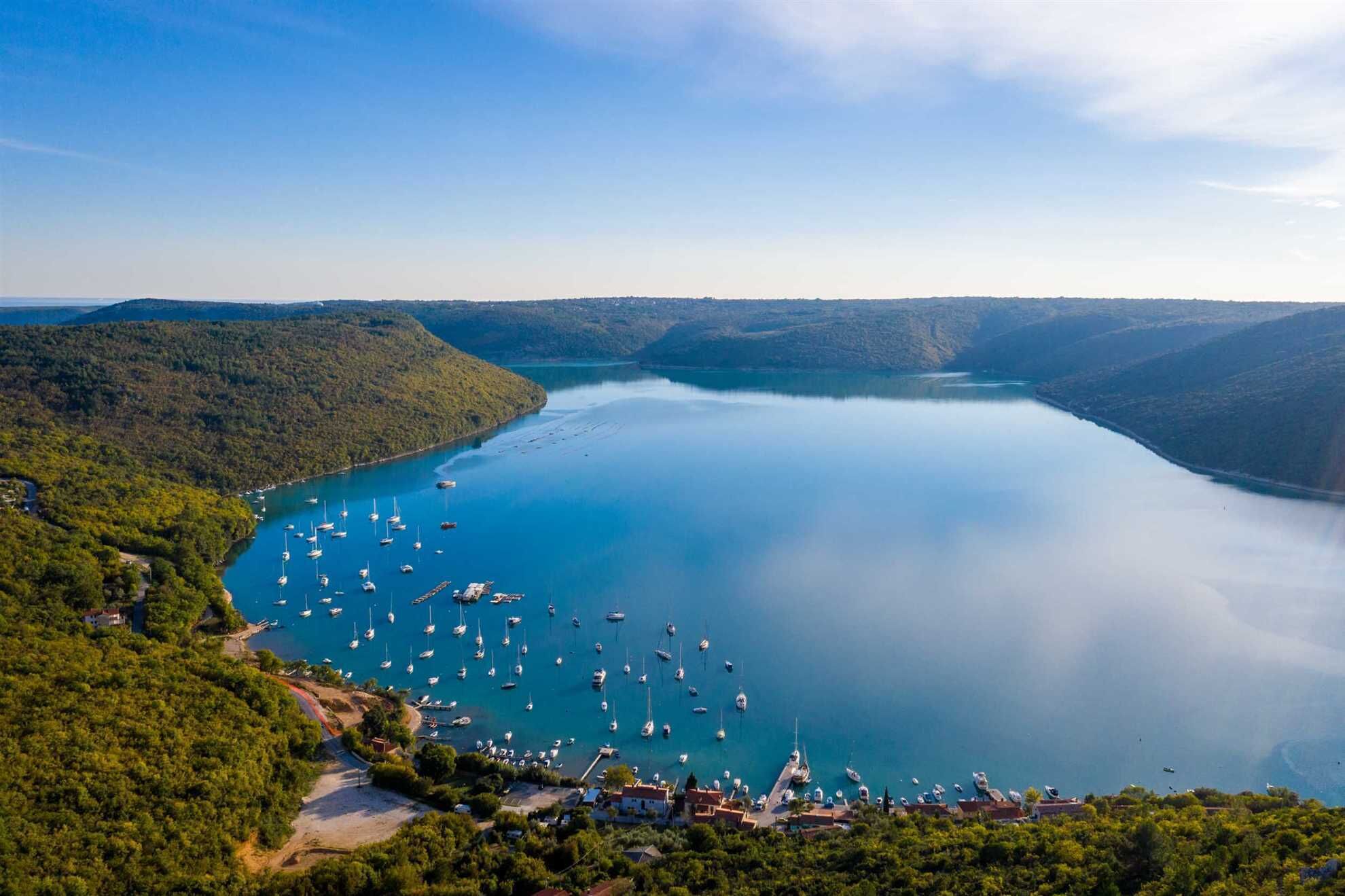 Une zone côtière dotée d’un port de plaisance rempli de bateaux, entourée de collines boisées.