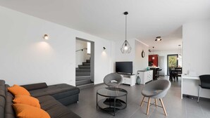 Una sala moderna con un sofá oscuro, una mesa de centro de vidrio y un televisor. Hay una escalera que lleva a otro nivel, una zona de cocina con una mesa de comedor y una silla.