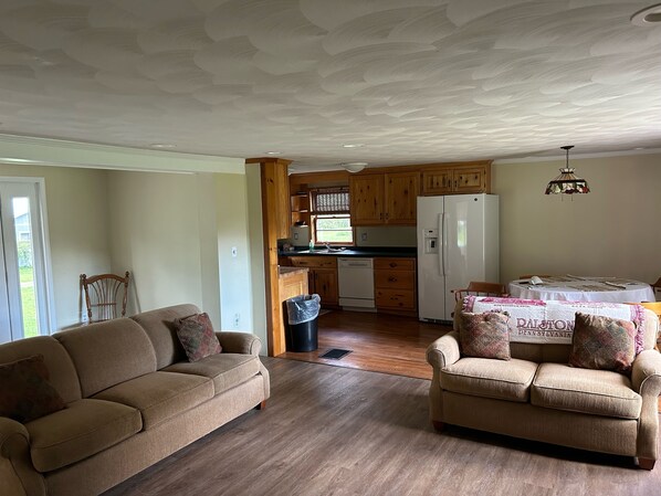 A cozy living room with two sofas, a dining table with a tablecloth, wooden cabinets, a white refrigerator, and a ceiling with a textured design.