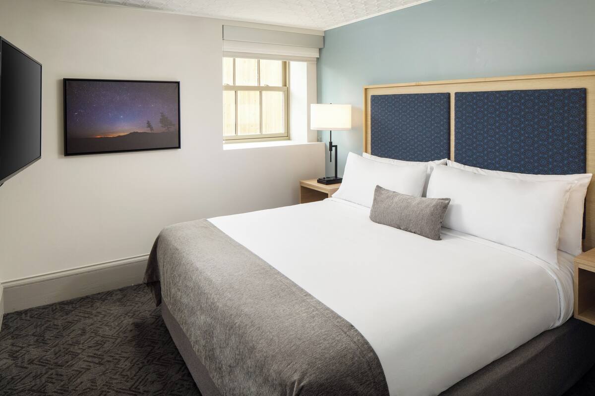 A neatly made bed with a grey throw blanket, a bedside table with a lamp, a framed picture of a starry night, and a window with a view of trees.