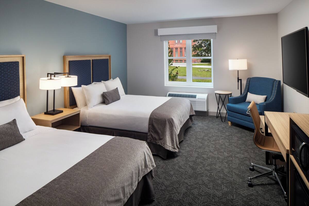 A hotel room with two beds, a desk, a chair, a TV, a window with a view of a building, and a lamp.