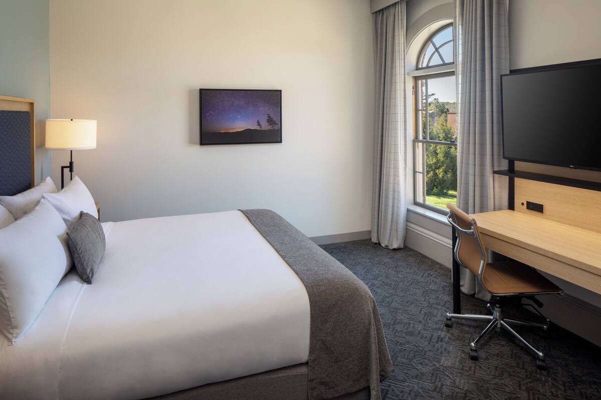 A hotel room with a large bed, a desk, a TV, a nightstand, and a window with curtains.