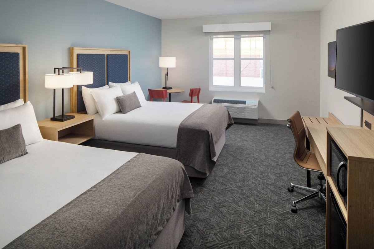 A hotel room with two beds, a desk, a chair, a TV, and a window.