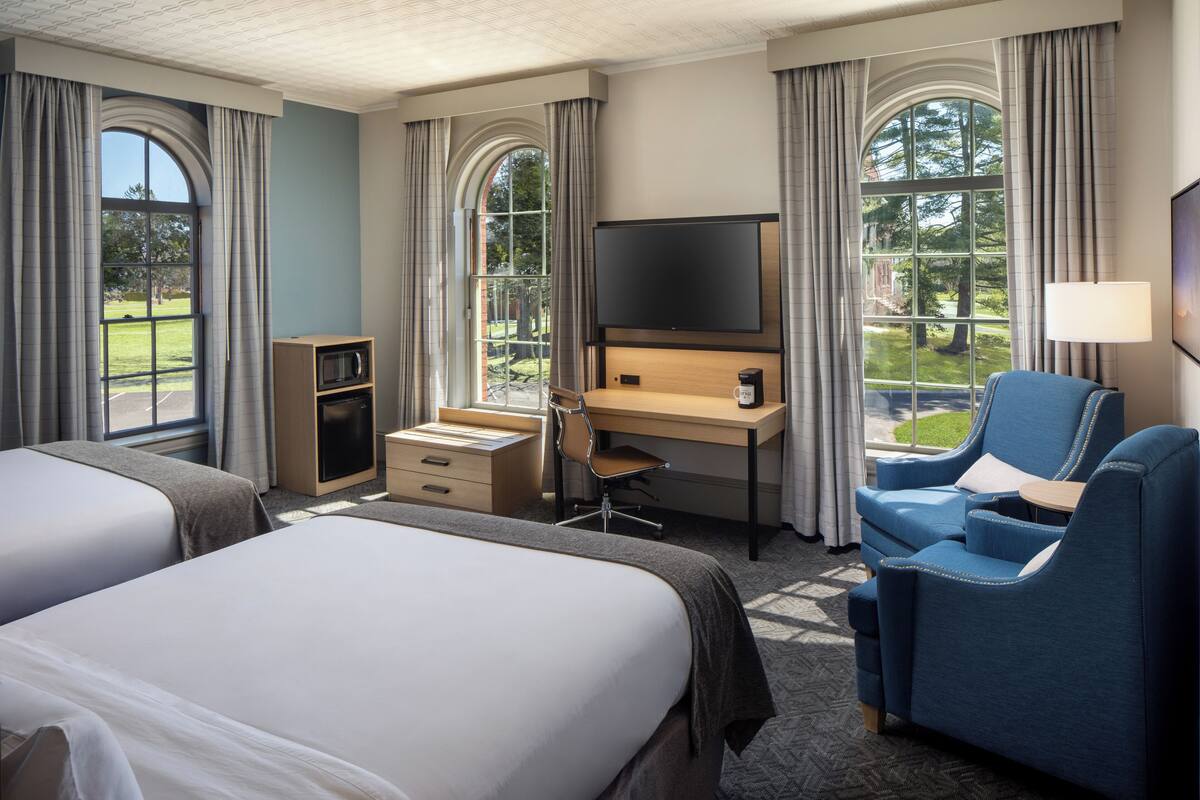 A hotel room with two beds, a desk, a chair, a TV, and large windows with curtains.