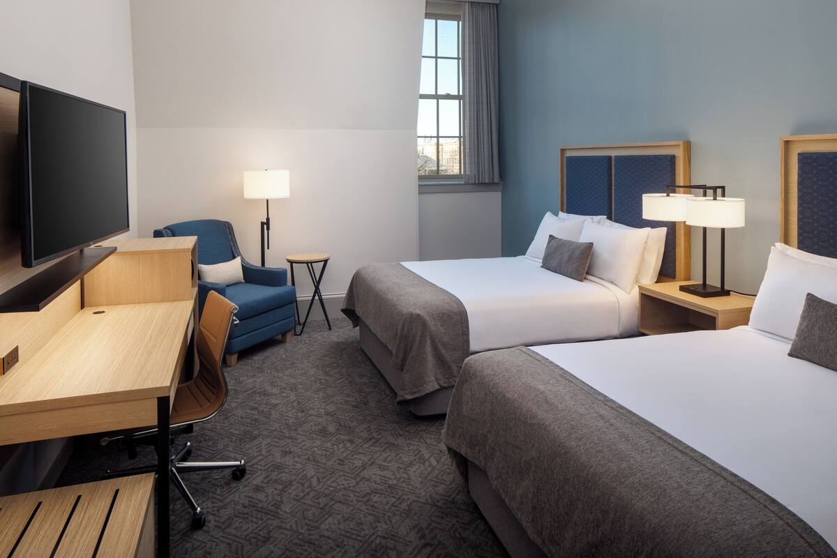 A hotel room with two beds, a desk, a chair, a TV, and a lamp.