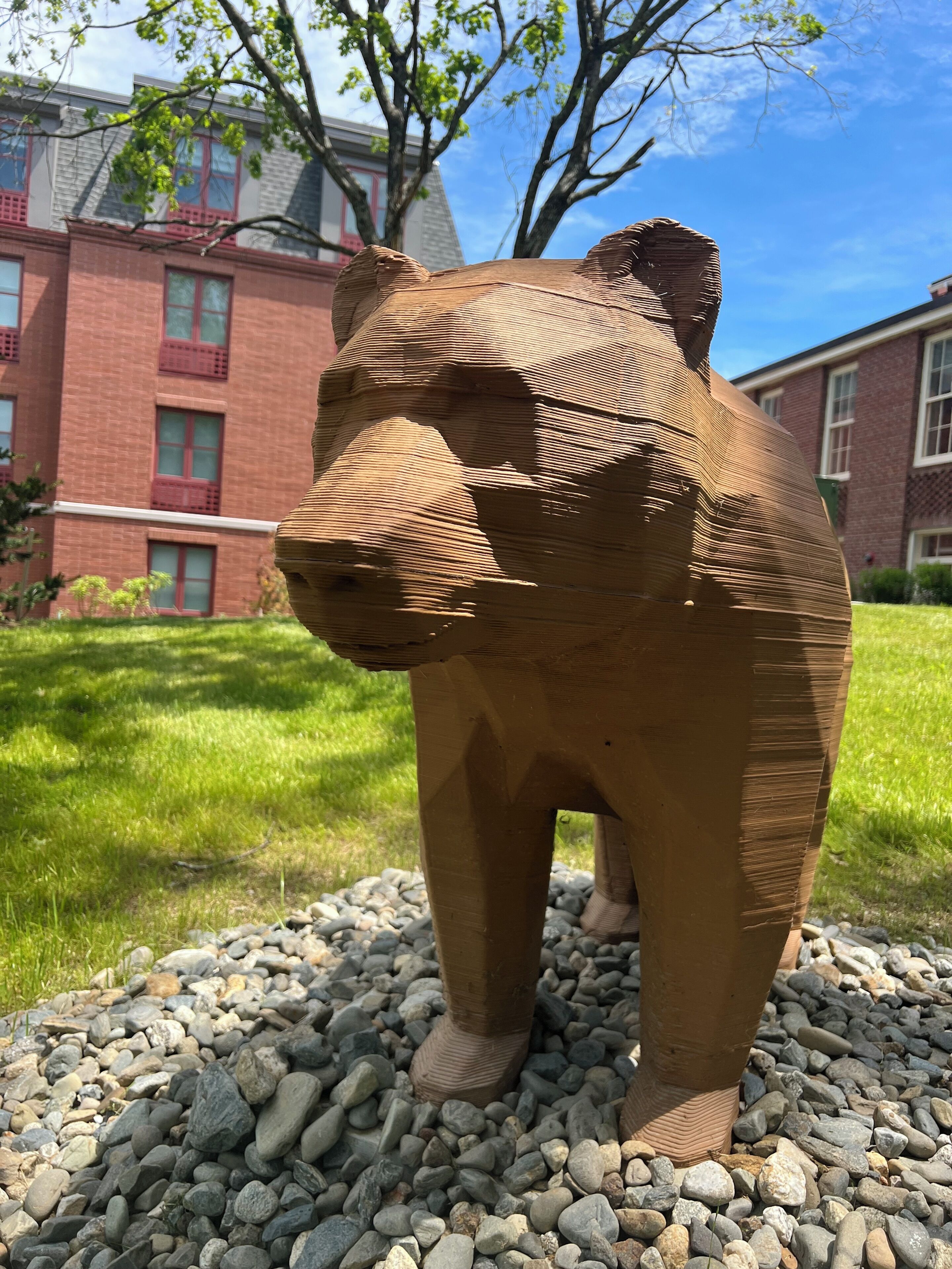 A sculpture of a bear made of bricks, standing on a bed of rocks in a grassy area with buildings in the background.