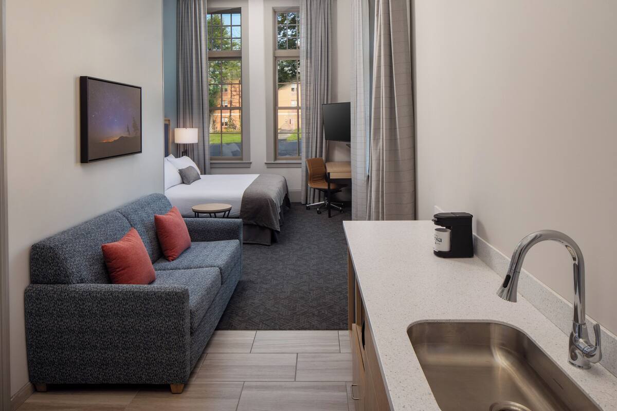 A modern hotel room with a sofa, bed, desk, and kitchenette.