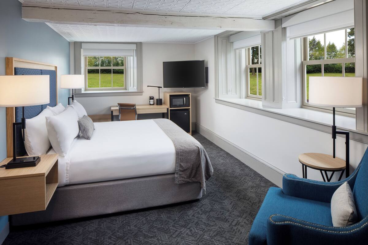 A hotel room with a bed, bedside tables, a desk with a TV, a chair, and large windows with a view of greenery.