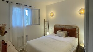 Une chambre à coucher avec un lit, une fenêtre avec des rideaux, une table de chevet et un luminaire fixé au mur.