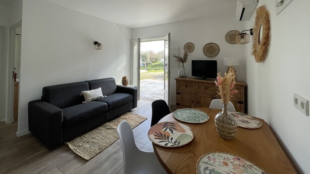 Un salon moderne avec un canapé foncé, une table à manger en bois agrémentée de sets de table, un téléviseur posé sur un meuble en bois, et une vue sur l’extérieur grâce à une porte vitrée.