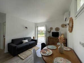 Ein modernes Wohnzimmer mit einer dunkelgrauen Couch, einem Holzesstisch mit Untersetzern, einem Fernseher auf einer Holzkommode und Blick ins Freie durch eine Glastür.