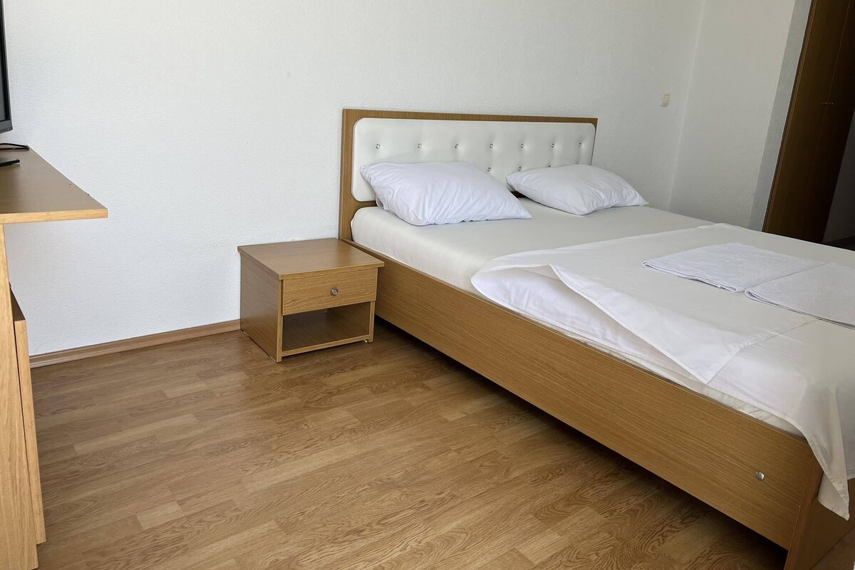 A single bed with a wooden frame and white bedding, a small wooden nightstand with a drawer, and a wooden desk with a chair.