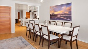 A modern dining area with a white table and wooden chairs, a kitchen in the background, and a cityscape painting on the wall. - So Close to Skiing at Sunny Ice House! Parking, Pool, Hot Tub (Telluride)