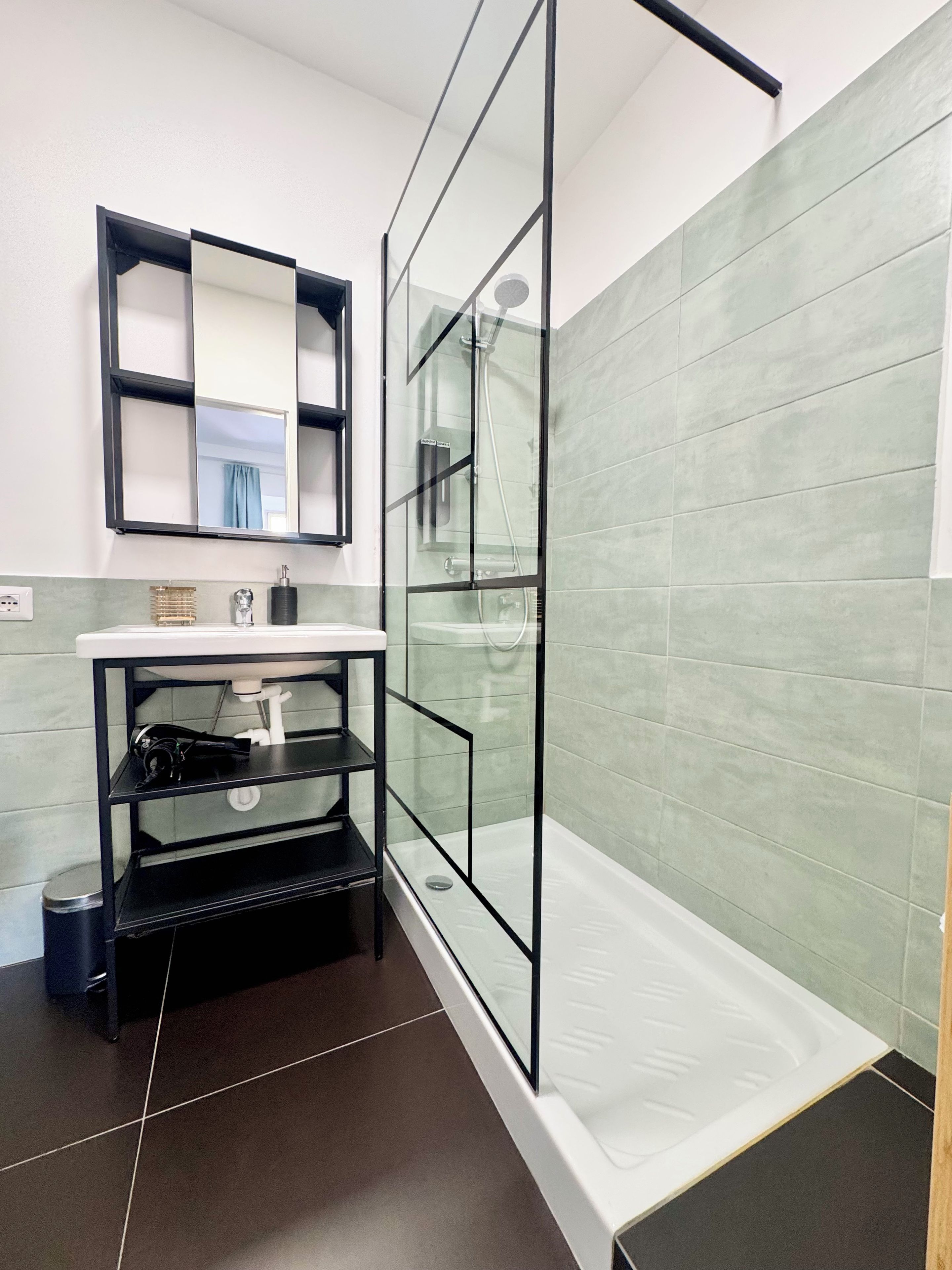 A modern bathroom with a glass shower enclosure, a white sink, and a black shelf unit.