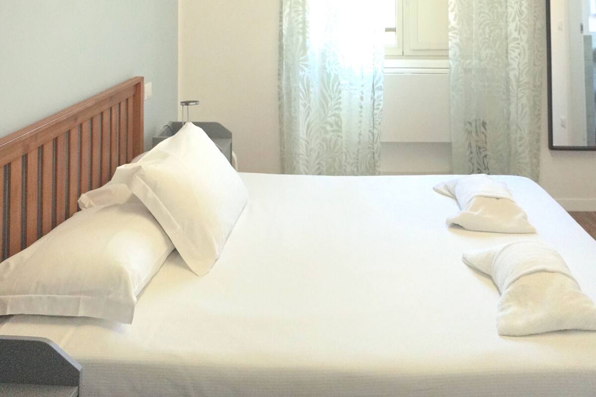 A hotel room with a neatly made bed, a wooden headboard, a nightstand, and a window with curtains.