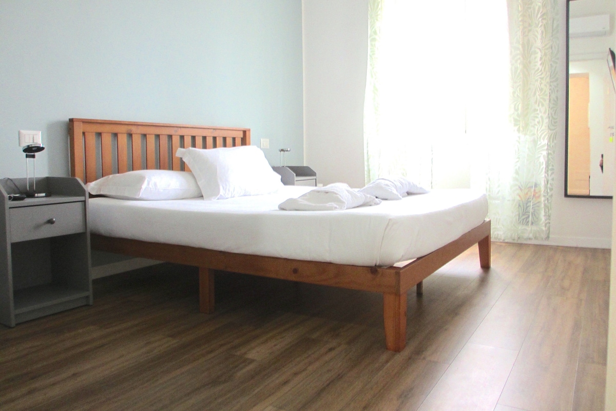 A neatly made bed with white linens in a room with wooden flooring, a nightstand, and a bedside table.